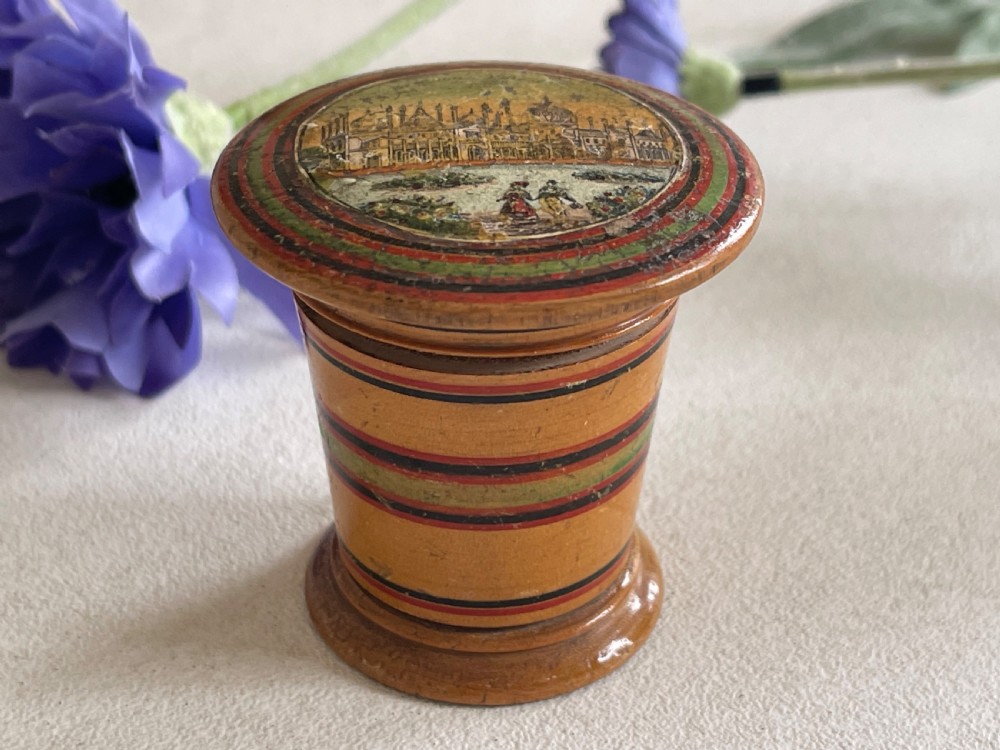 A small turned wooden Georgian nutmeg grater featuring painted red and green stripes around its cylindrical body and a circular printed landscape scene on the lid.