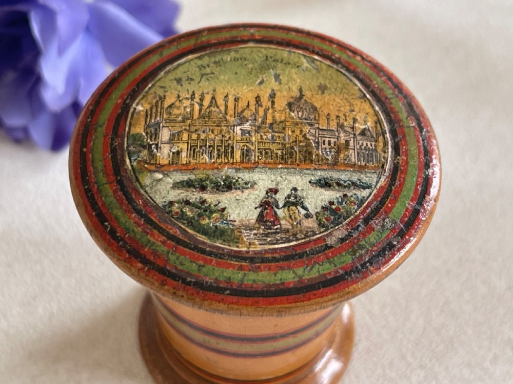 A small turned wooden Georgian nutmeg grater featuring painted red and green stripes around its cylindrical body and a circular printed landscape scene on the lid. View II