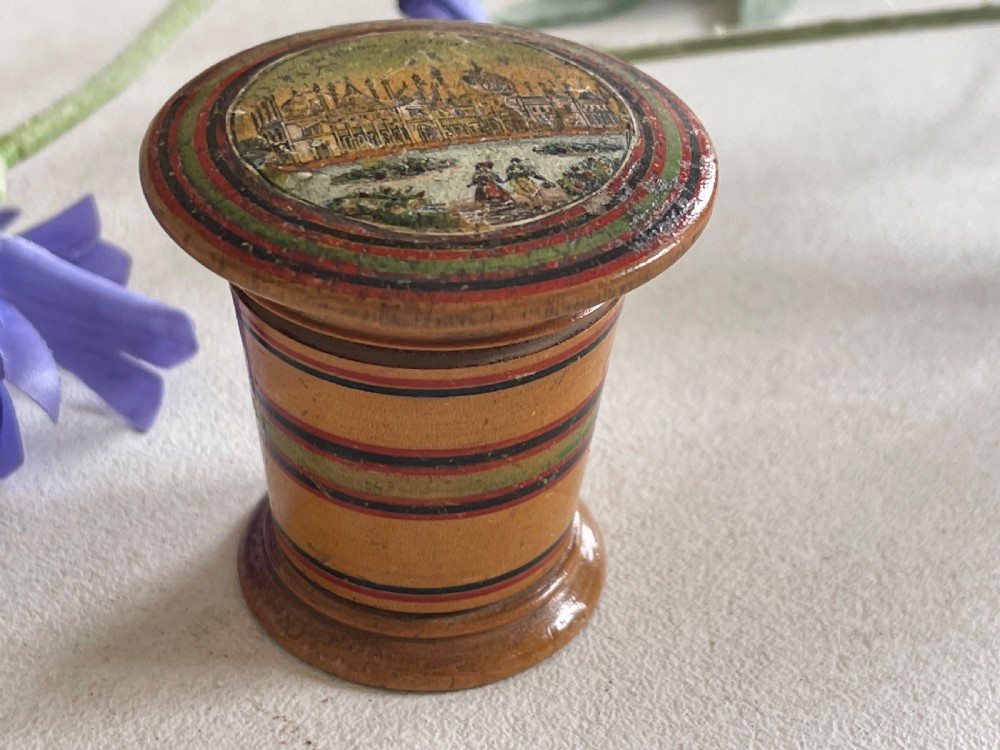 A small turned wooden Georgian nutmeg grater featuring painted red and green stripes around its cylindrical body and a circular printed landscape scene on the lid. View V
