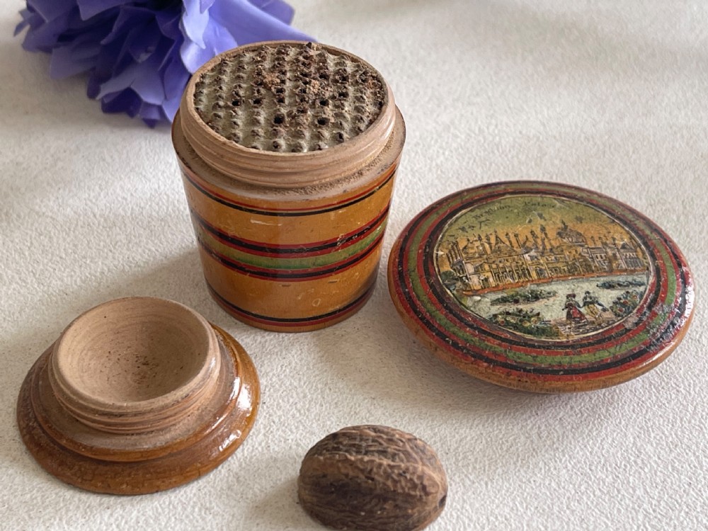 A small turned wooden Georgian nutmeg grater featuring painted red and green stripes around its cylindrical body and a circular printed landscape scene on the lid. View VII