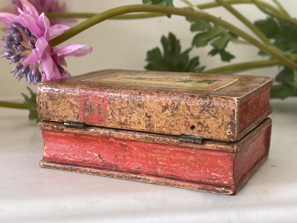 A small rectangular antique box constructed in the shape of two stacked books with red and gold patterned covers and exterior brass hinges.
