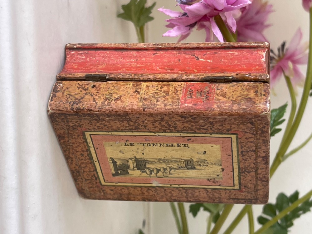 A small rectangular antique box constructed in the shape of two stacked books with red and gold patterned covers and exterior brass hinges. View II
