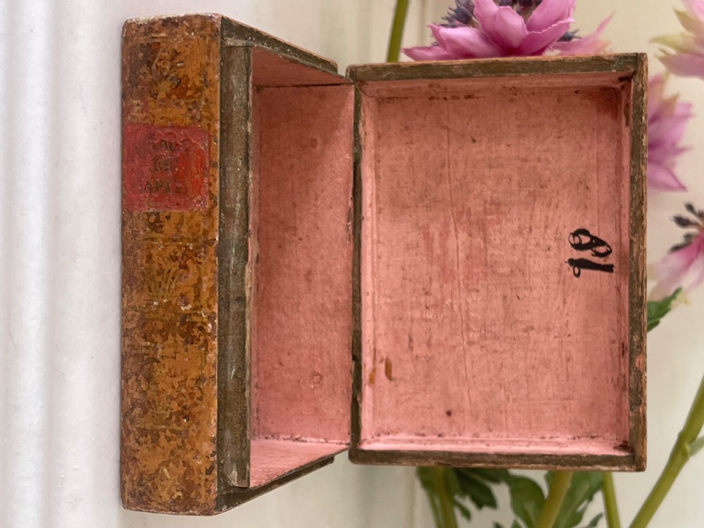 A small rectangular antique box constructed in the shape of two stacked books with red and gold patterned covers and exterior brass hinges. View III