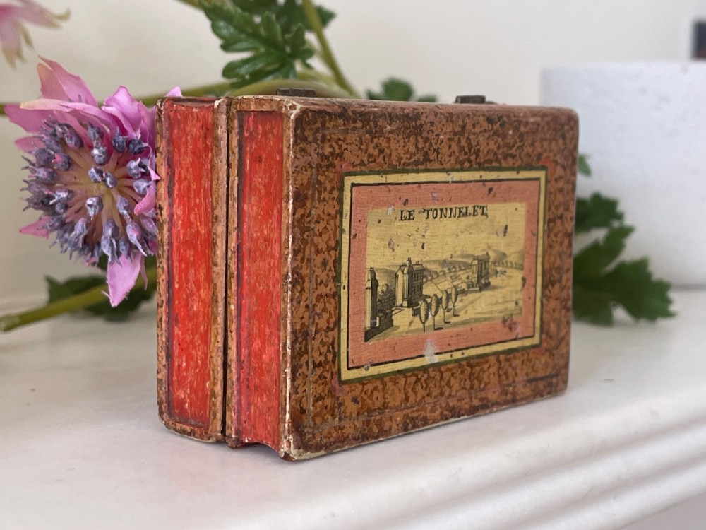 A small rectangular antique box constructed in the shape of two stacked books with red and gold patterned covers and exterior brass hinges. View V