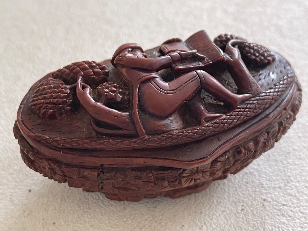 An oval-shaped dark brown coquilla nut snuff box featuring an intricate high-relief carved scene of a seated figure with a writing desk and stylized trees. View III