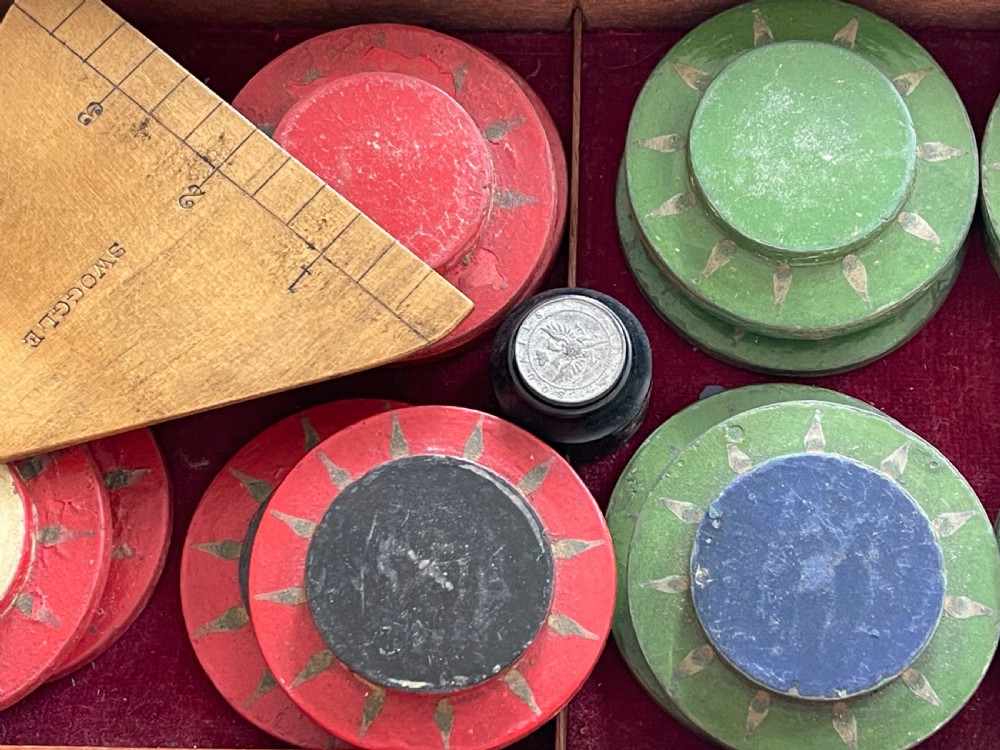 An open wooden mahogany box containing a Victorian squails game set with colourful turned wooden discs and a triangular wooden swoggle measuring tool. View IV