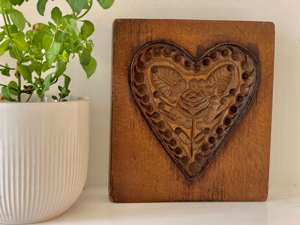 A square wooden block featuring a deeply carved heart motif with a central rose, leaves, and patterned borders outlined by embedded metal.