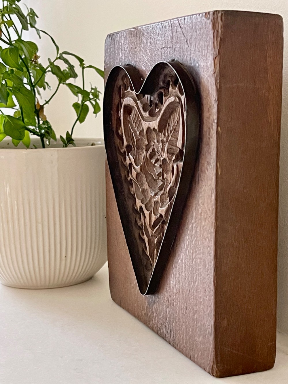 A square wooden block featuring a deeply carved heart motif with a central rose, leaves, and patterned borders outlined by embedded metal. View II