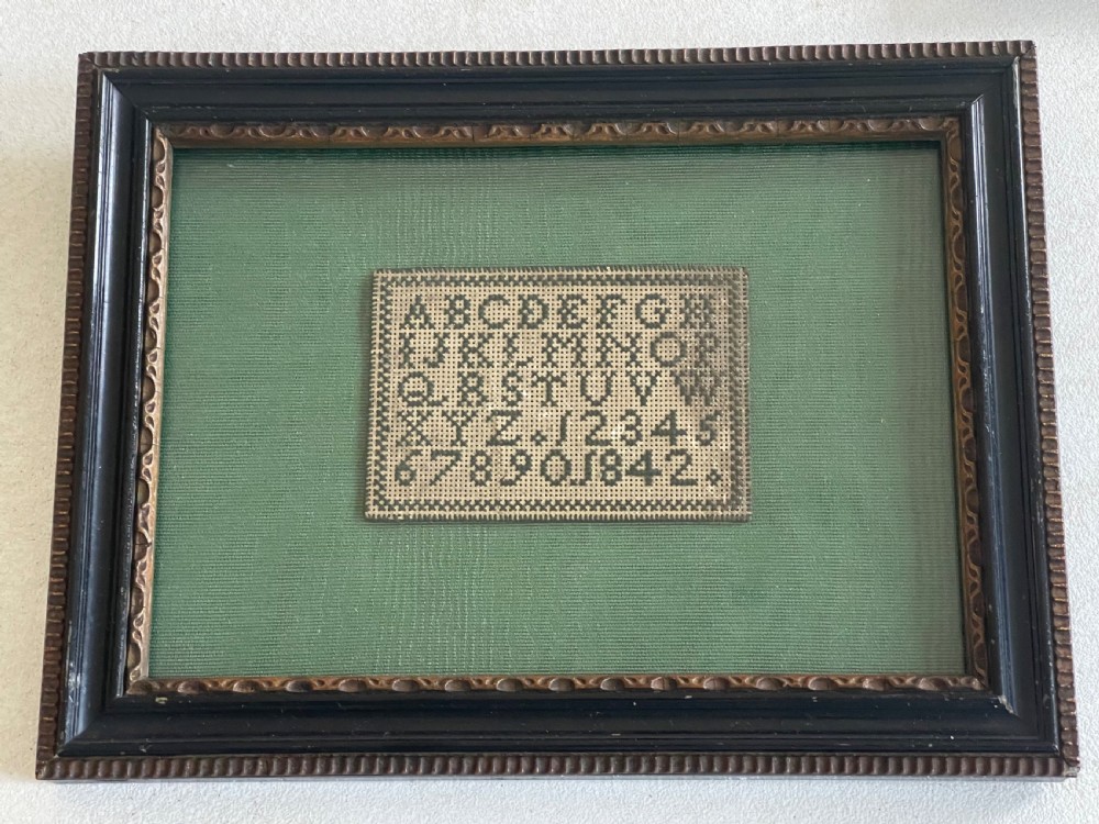 A framed Victorian miniature needlework sampler displaying the alphabet and numbers mounted on green fabric within a dark carved wooden frame.