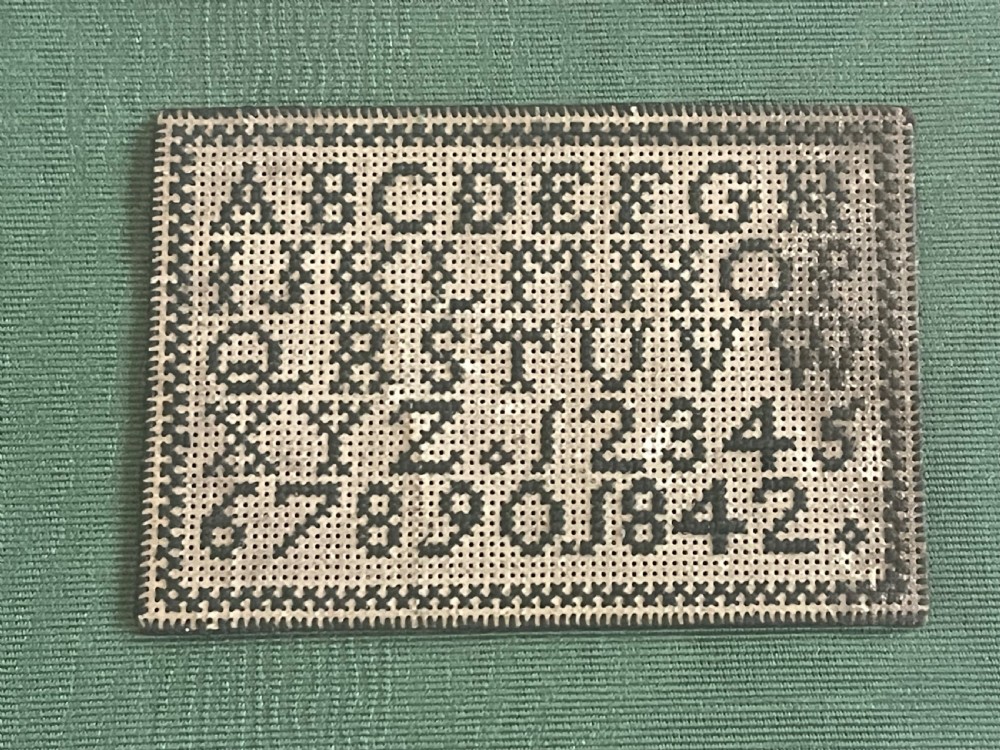 A framed Victorian miniature needlework sampler displaying the alphabet and numbers mounted on green fabric within a dark carved wooden frame. View II