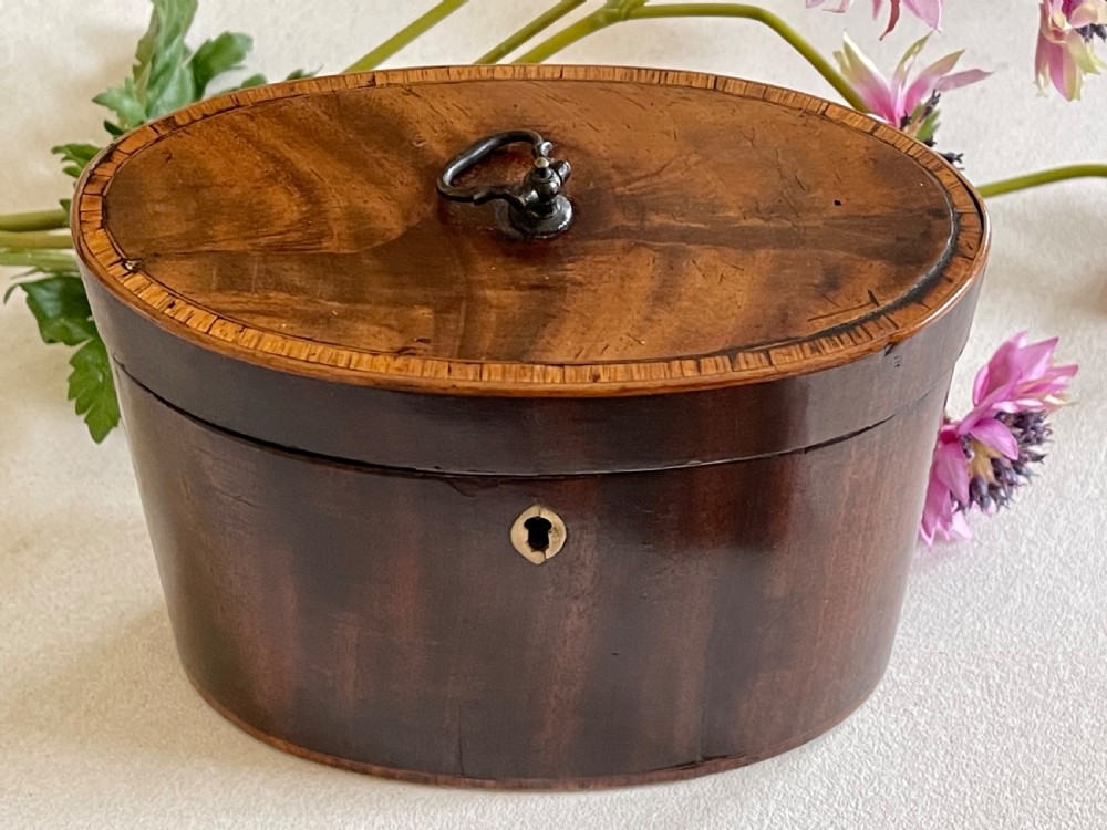 An eighteenth-century Georgian oval mahogany tea caddy featuring polished grain, a bone keyhole escutcheon, and a brass ring handle on the hinged top. View V