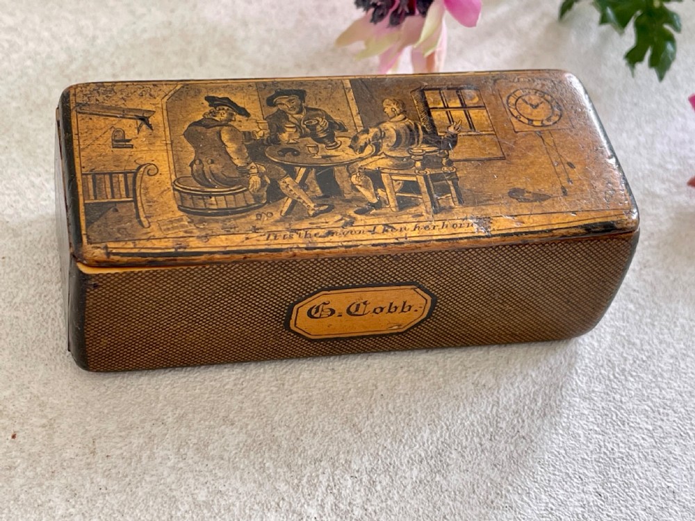 A small rectangular antique wooden snuff box featuring penwork illustrations of men in a tavern on the lid and a central name tag inscribed G. Cobb on the front.