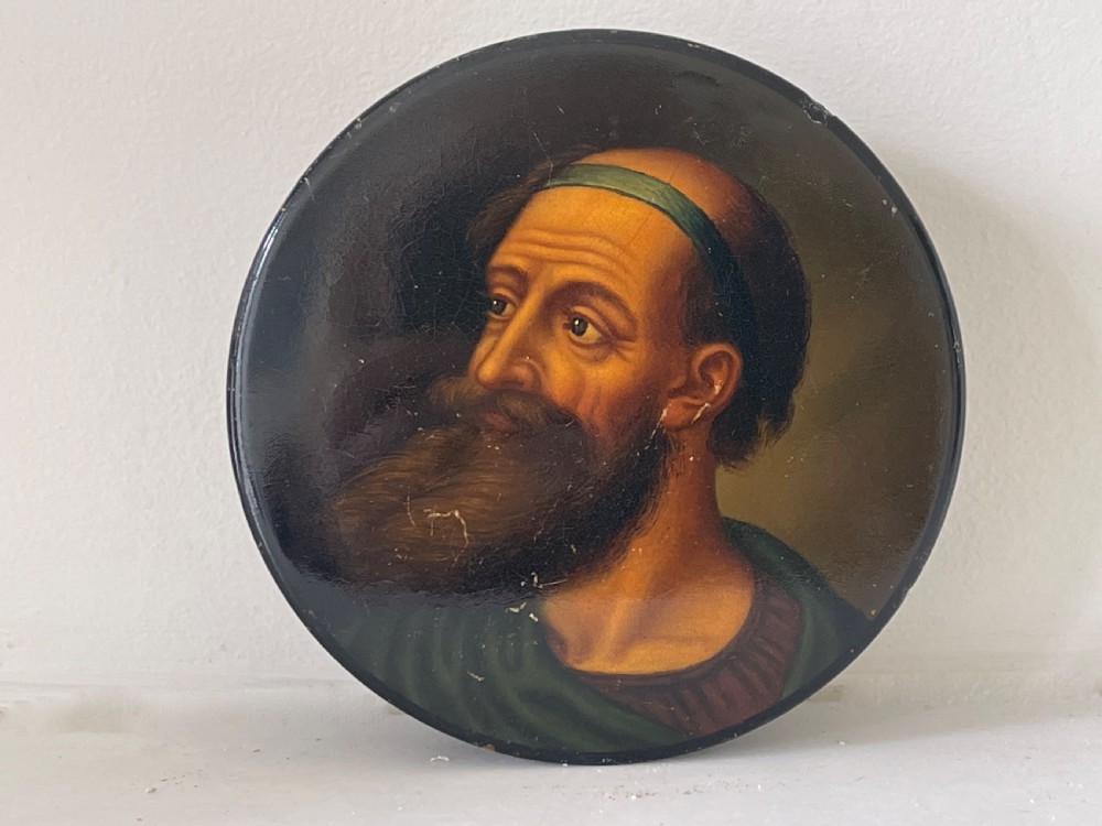 A circular black lacquered papier-mache snuff box lid featuring a hand-painted classical portrait of a bearded man wearing a green headband.