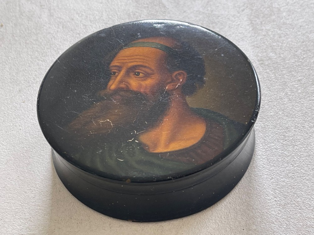 A circular black lacquered papier-mache snuff box lid featuring a hand-painted classical portrait of a bearded man wearing a green headband. View V