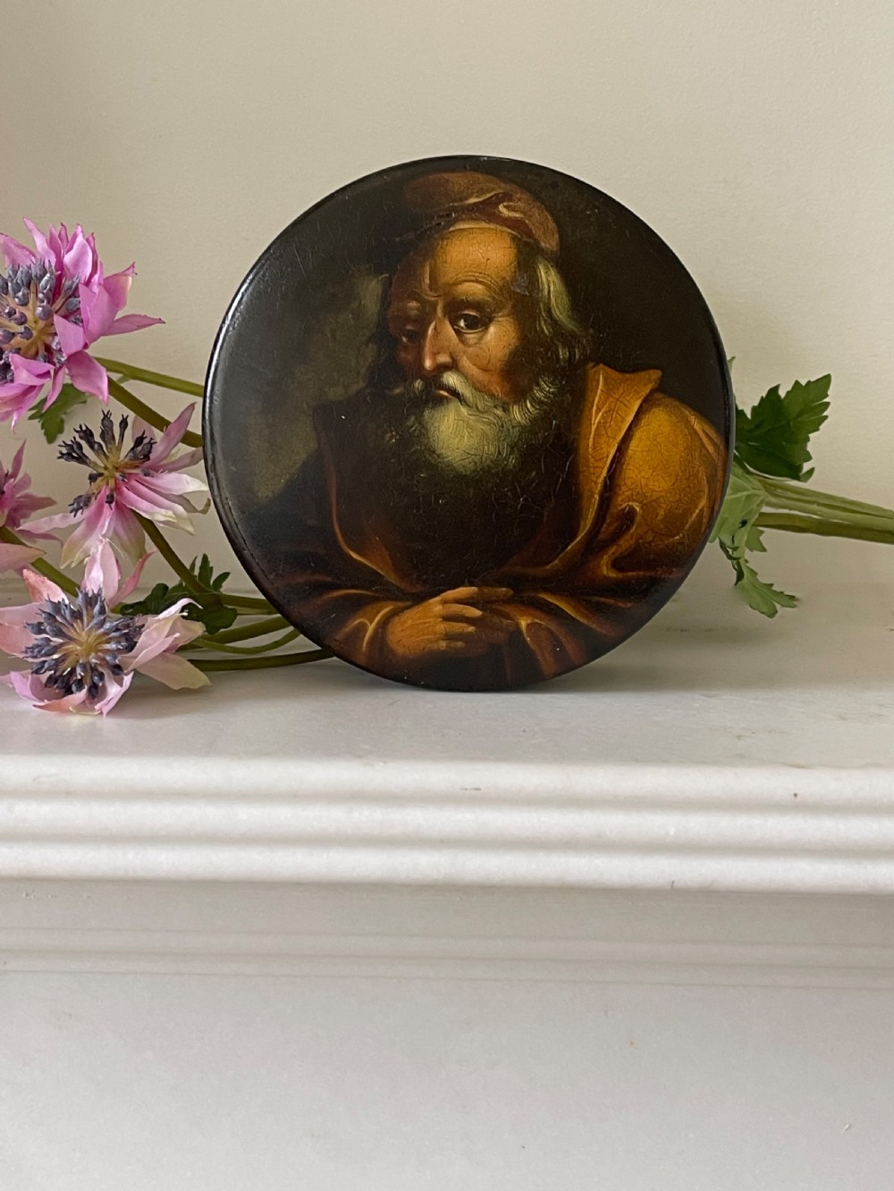 A circular dark brown antique papier-mache snuff box featuring a detailed painted miniature portrait of a bearded man on the lid.