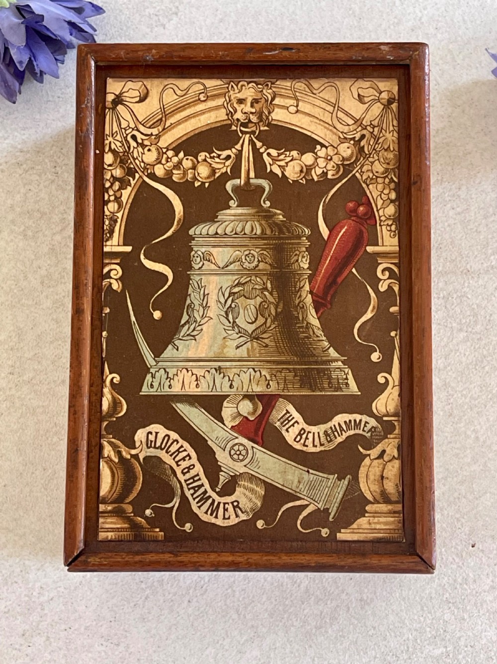 A rectangular wooden game box with a detailed printed paper lid depicting a bell, a hammer, floral garlands, and classical scrollwork.