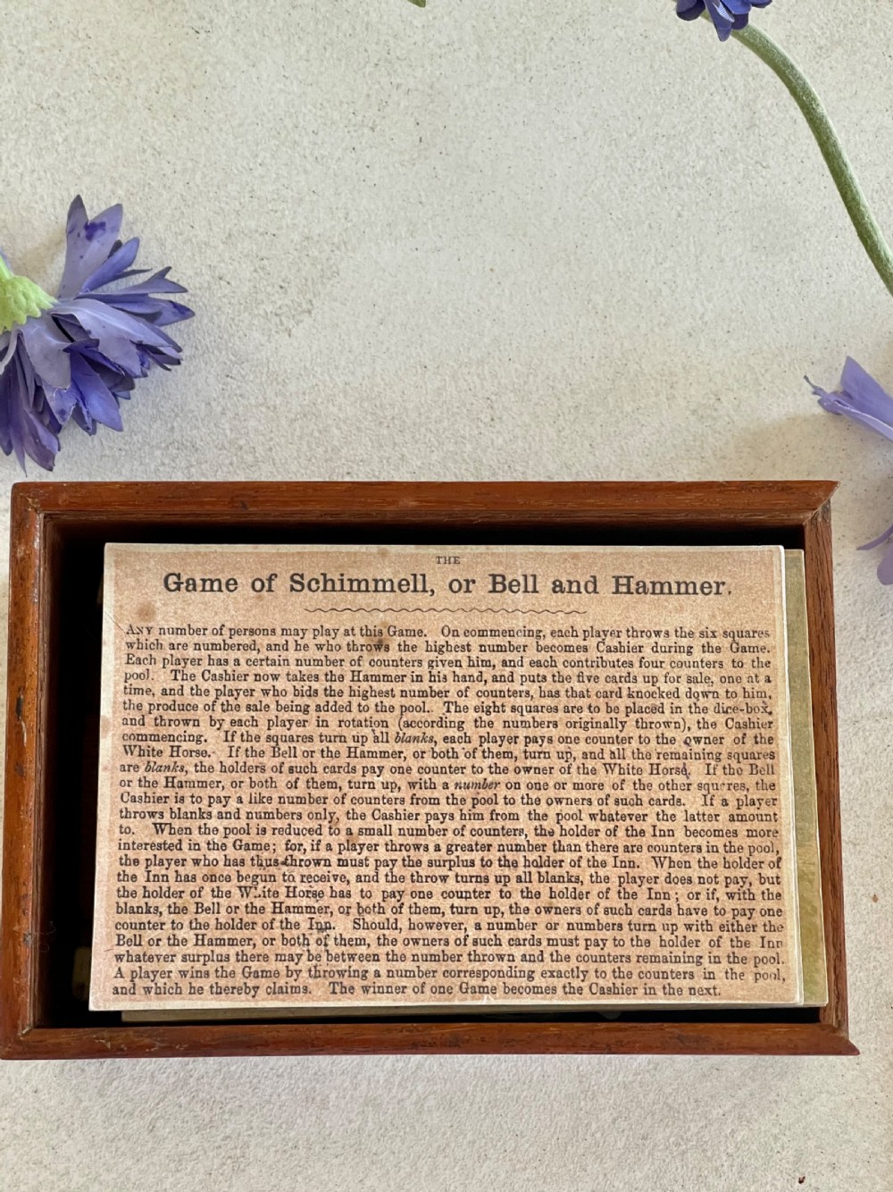 A rectangular wooden game box with a detailed printed paper lid depicting a bell, a hammer, floral garlands, and classical scrollwork. View VI
