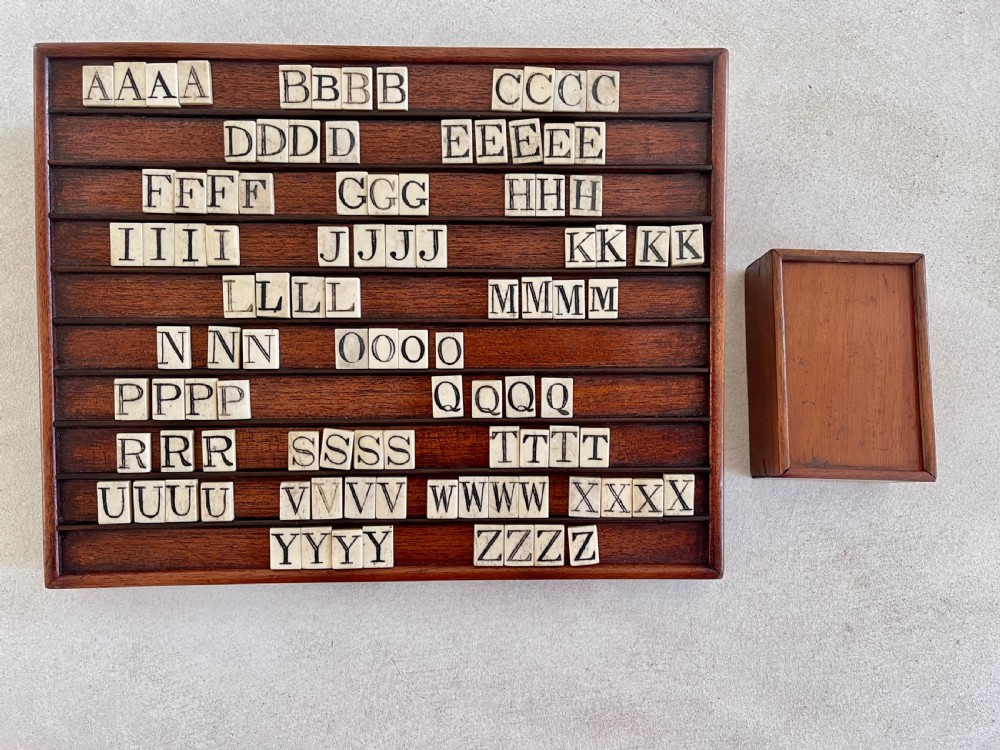 A dark mahogany wooden alphabet slope featuring horizontal ledges holding square carved bone tiles engraved with capital letters, beside a small rectangular wooden storage box.