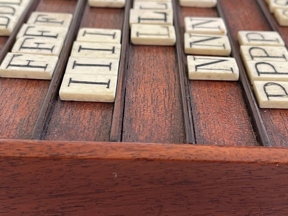 A dark mahogany wooden alphabet slope featuring horizontal ledges holding square carved bone tiles engraved with capital letters, beside a small rectangular wooden storage box. View III