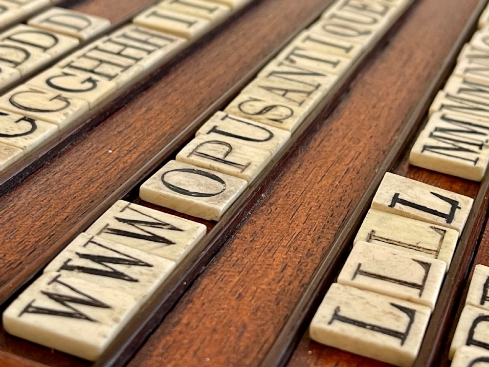 A dark mahogany wooden alphabet slope featuring horizontal ledges holding square carved bone tiles engraved with capital letters, beside a small rectangular wooden storage box. View IV