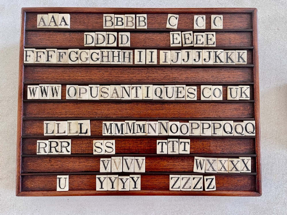 A dark mahogany wooden alphabet slope featuring horizontal ledges holding square carved bone tiles engraved with capital letters, beside a small rectangular wooden storage box. View V