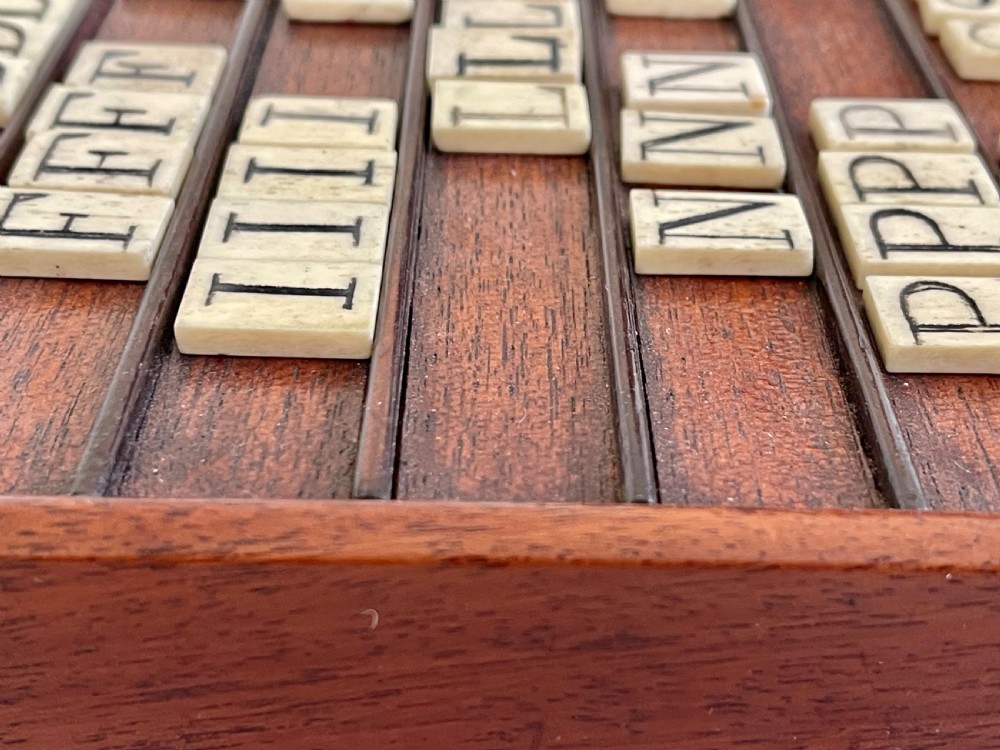 A dark mahogany wooden alphabet slope featuring horizontal ledges holding square carved bone tiles engraved with capital letters, beside a small rectangular wooden storage box. View VI