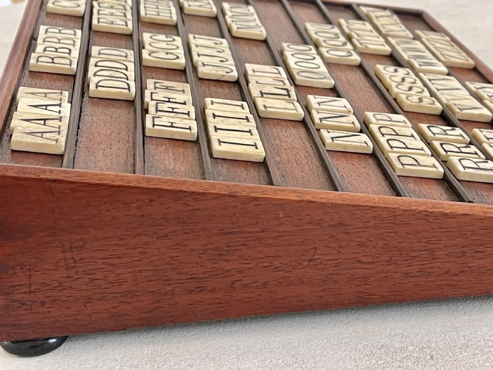 A dark mahogany wooden alphabet slope featuring horizontal ledges holding square carved bone tiles engraved with capital letters, beside a small rectangular wooden storage box. View VII