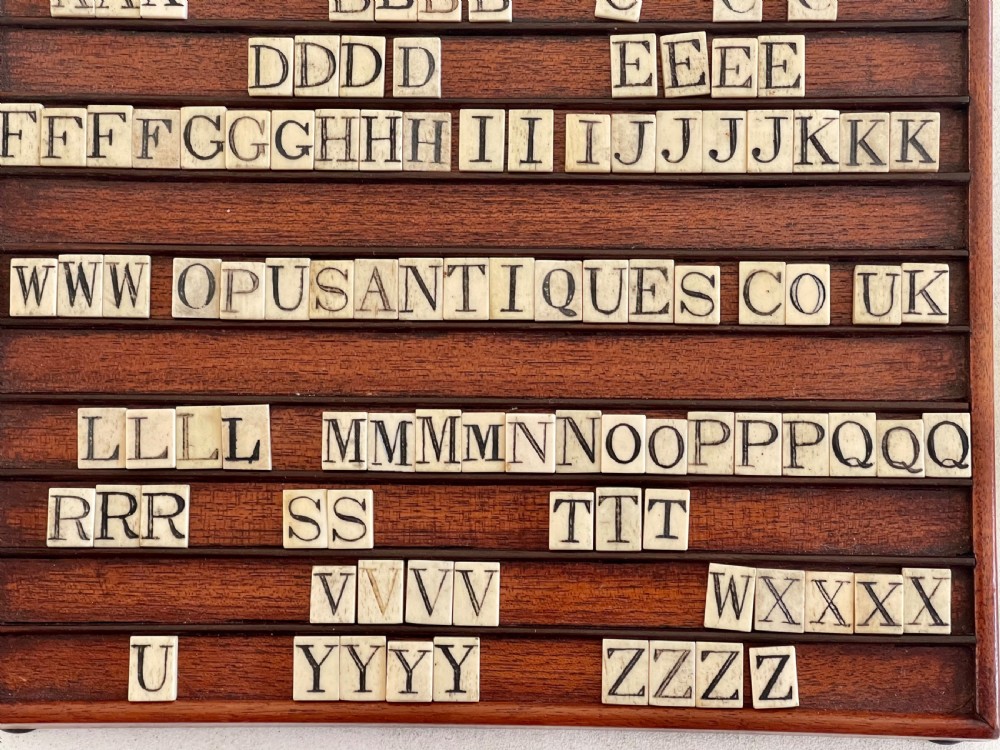 A dark mahogany wooden alphabet slope featuring horizontal ledges holding square carved bone tiles engraved with capital letters, beside a small rectangular wooden storage box. View VIII