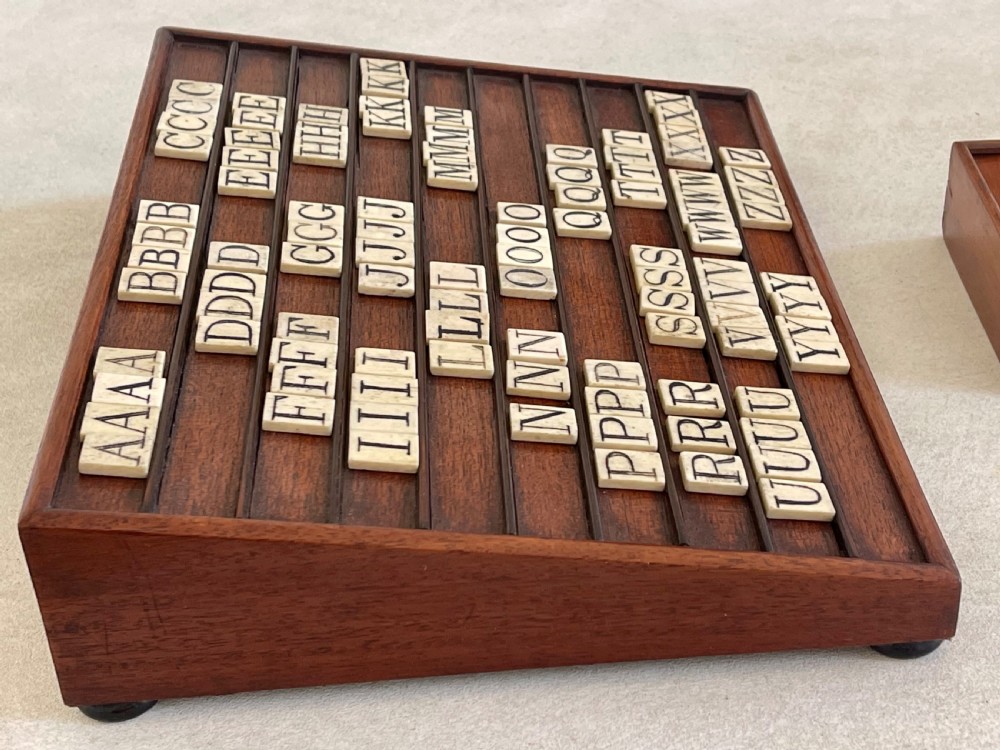 A dark mahogany wooden alphabet slope featuring horizontal ledges holding square carved bone tiles engraved with capital letters, beside a small rectangular wooden storage box. View IX