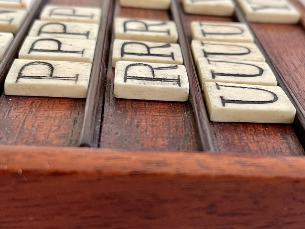 A dark mahogany wooden alphabet slope featuring horizontal ledges holding square carved bone tiles engraved with capital letters, beside a small rectangular wooden storage box. View X