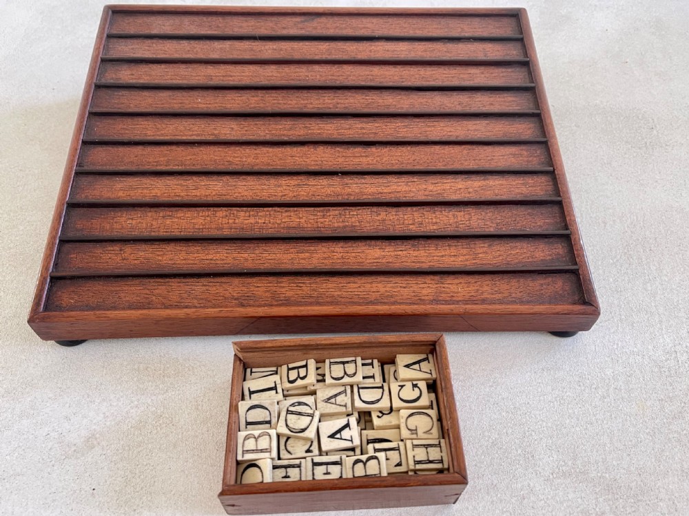 A dark mahogany wooden alphabet slope featuring horizontal ledges holding square carved bone tiles engraved with capital letters, beside a small rectangular wooden storage box. View XI
