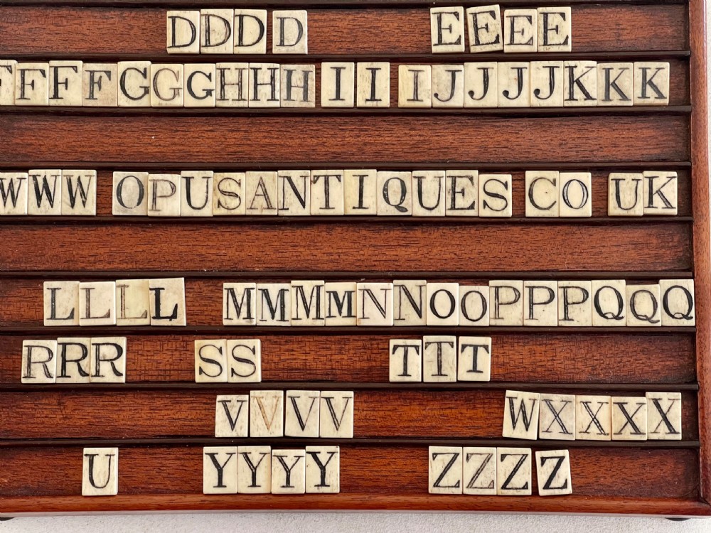 A dark mahogany wooden alphabet slope featuring horizontal ledges holding square carved bone tiles engraved with capital letters, beside a small rectangular wooden storage box. View XII
