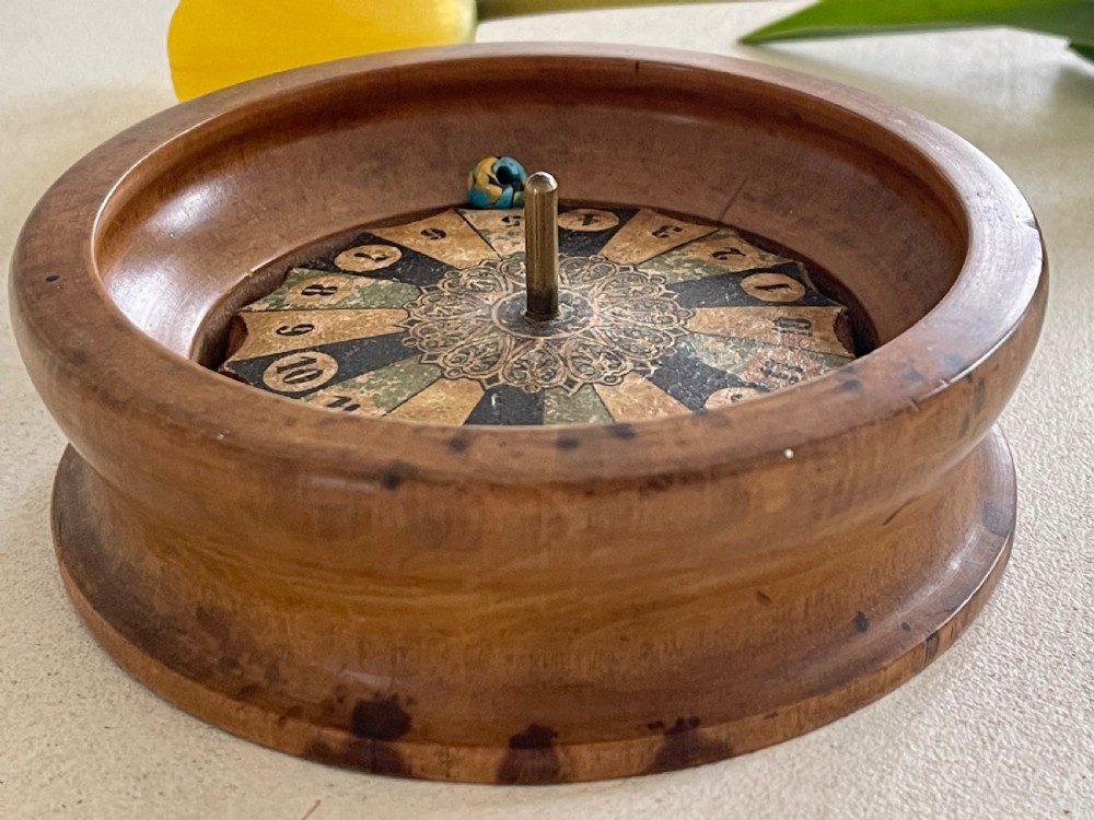 A turned wooden antique tabletop roulette wheel with a central brass spindle, printed numbered dial, and small marble ball.