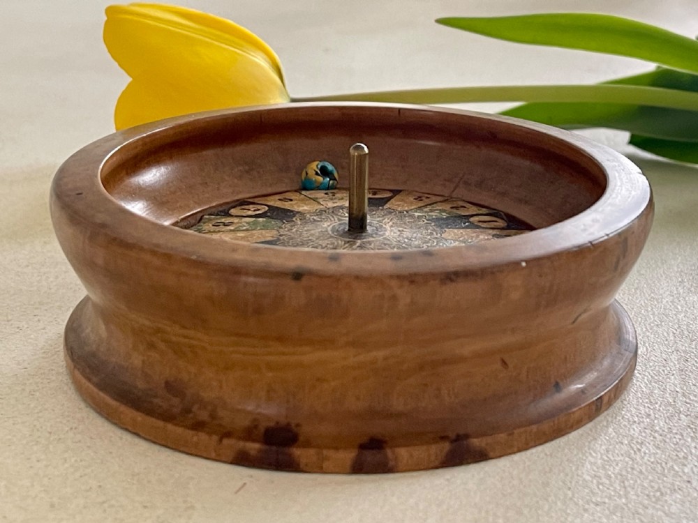 A turned wooden antique tabletop roulette wheel with a central brass spindle, printed numbered dial, and small marble ball. View III