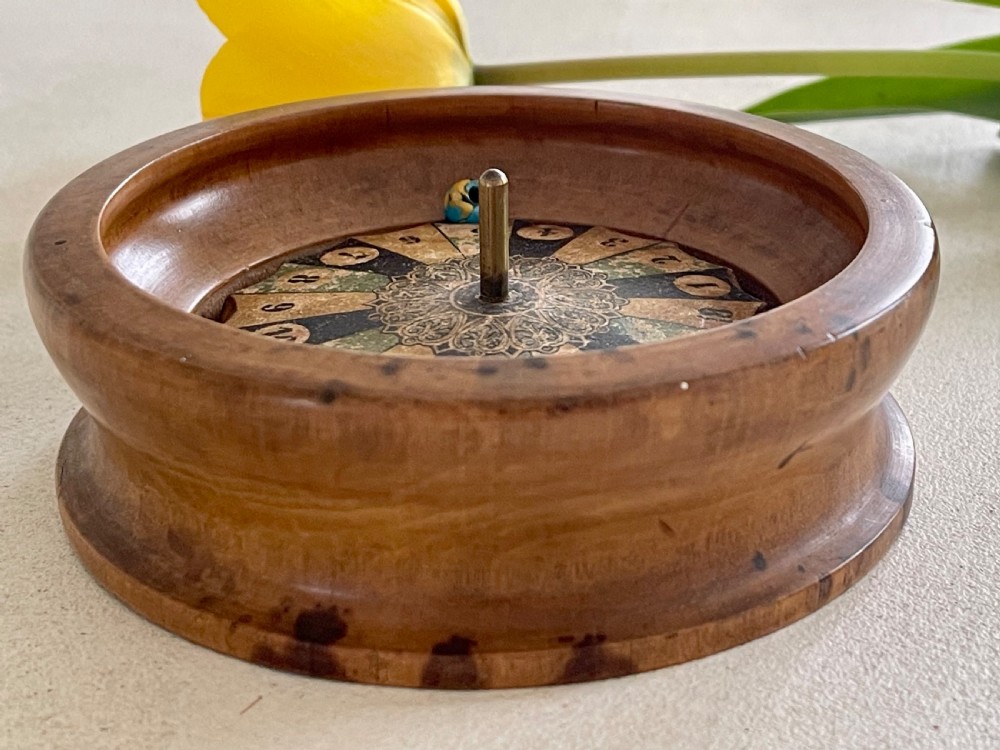 A turned wooden antique tabletop roulette wheel with a central brass spindle, printed numbered dial, and small marble ball. View IV