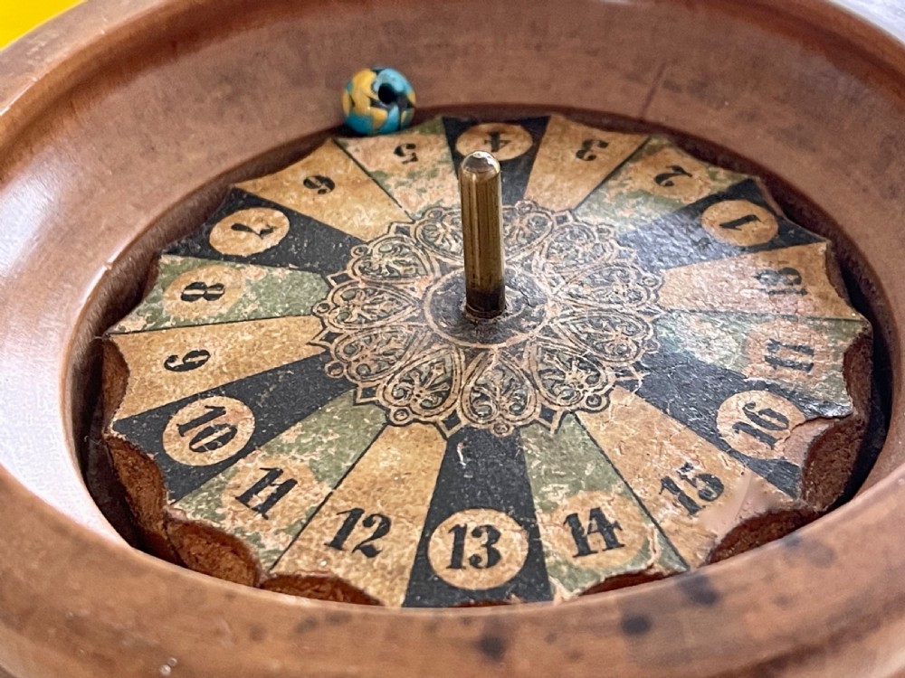 A turned wooden antique tabletop roulette wheel with a central brass spindle, printed numbered dial, and small marble ball. View V