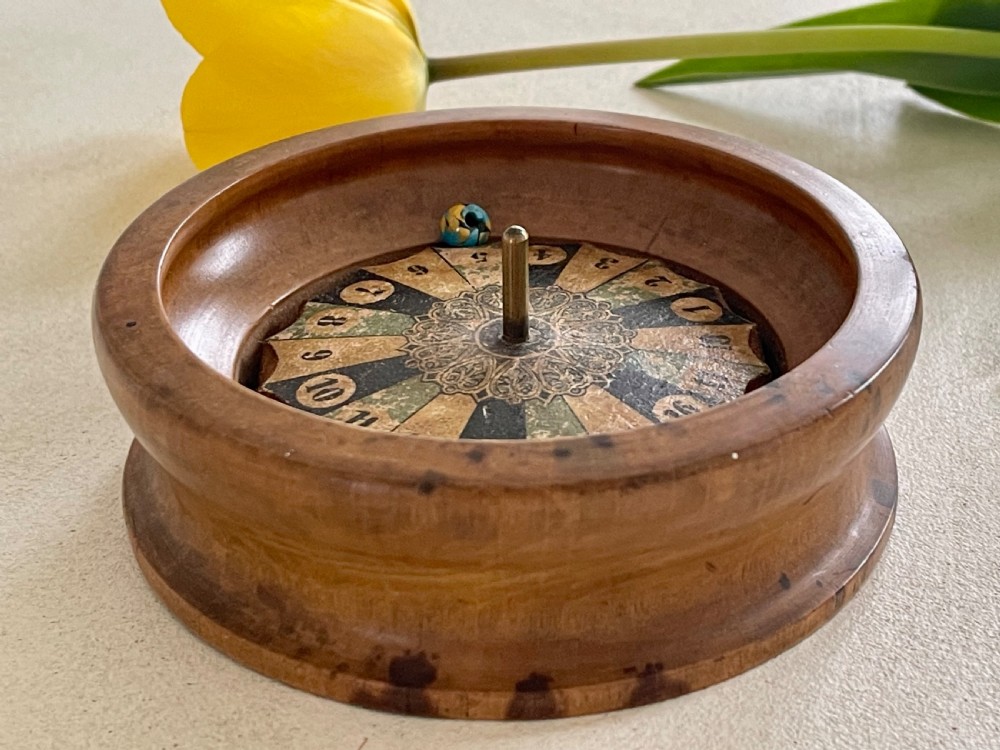 A turned wooden antique tabletop roulette wheel with a central brass spindle, printed numbered dial, and small marble ball. View VI