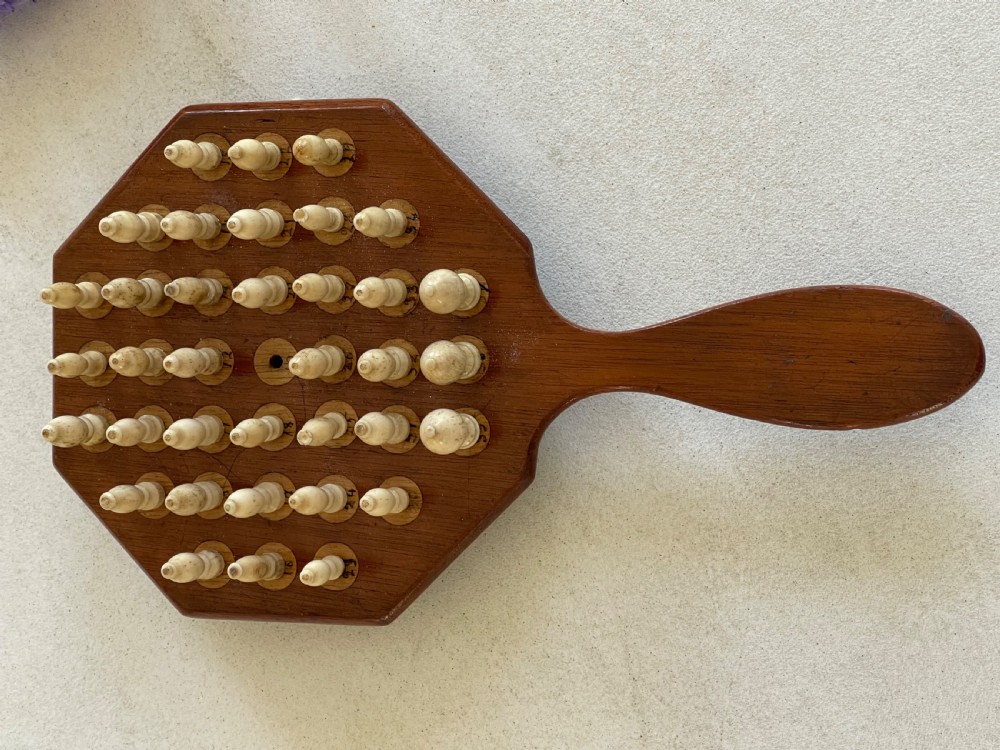 A paddle-shaped wooden peg solitaire board featuring numbered slots and carved bone pegs. View VI