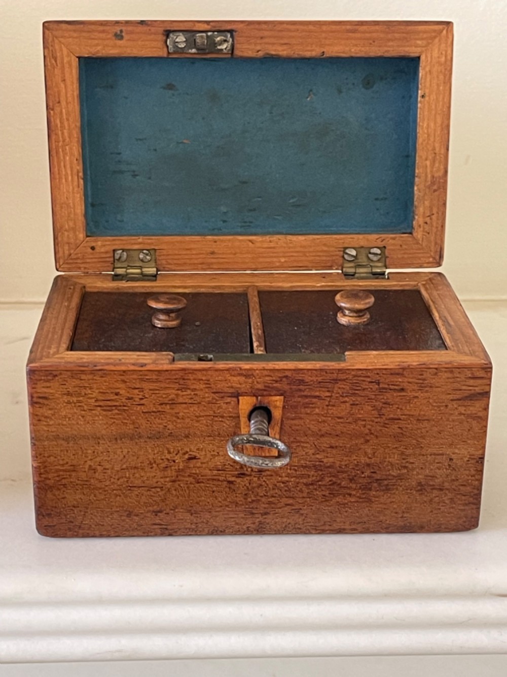 An open small wooden tea caddy featuring two internal compartments with turned knobs, a blue-lined interior lid, and a key inserted into the front lock.