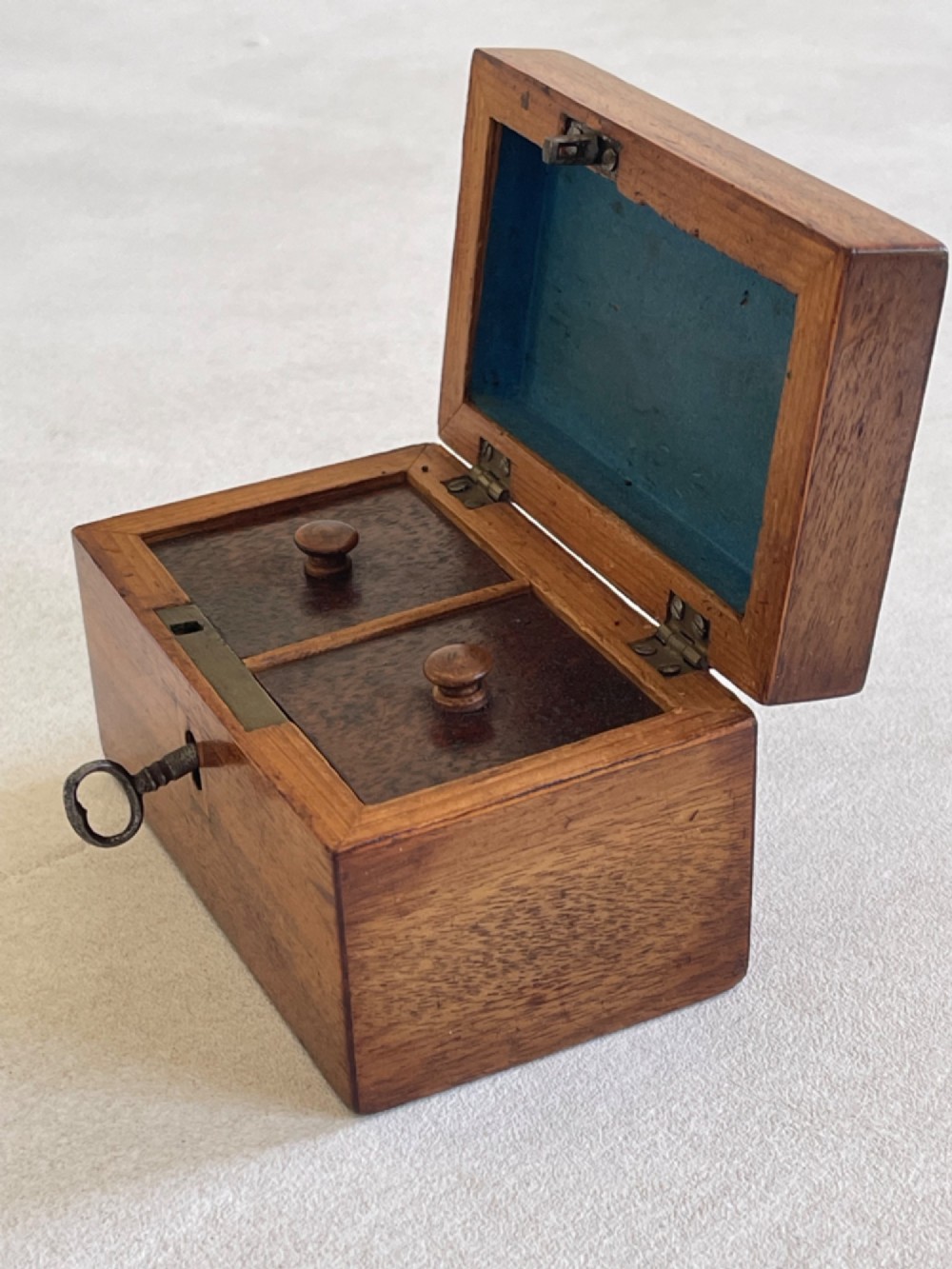 An open small wooden tea caddy featuring two internal compartments with turned knobs, a blue-lined interior lid, and a key inserted into the front lock. View III