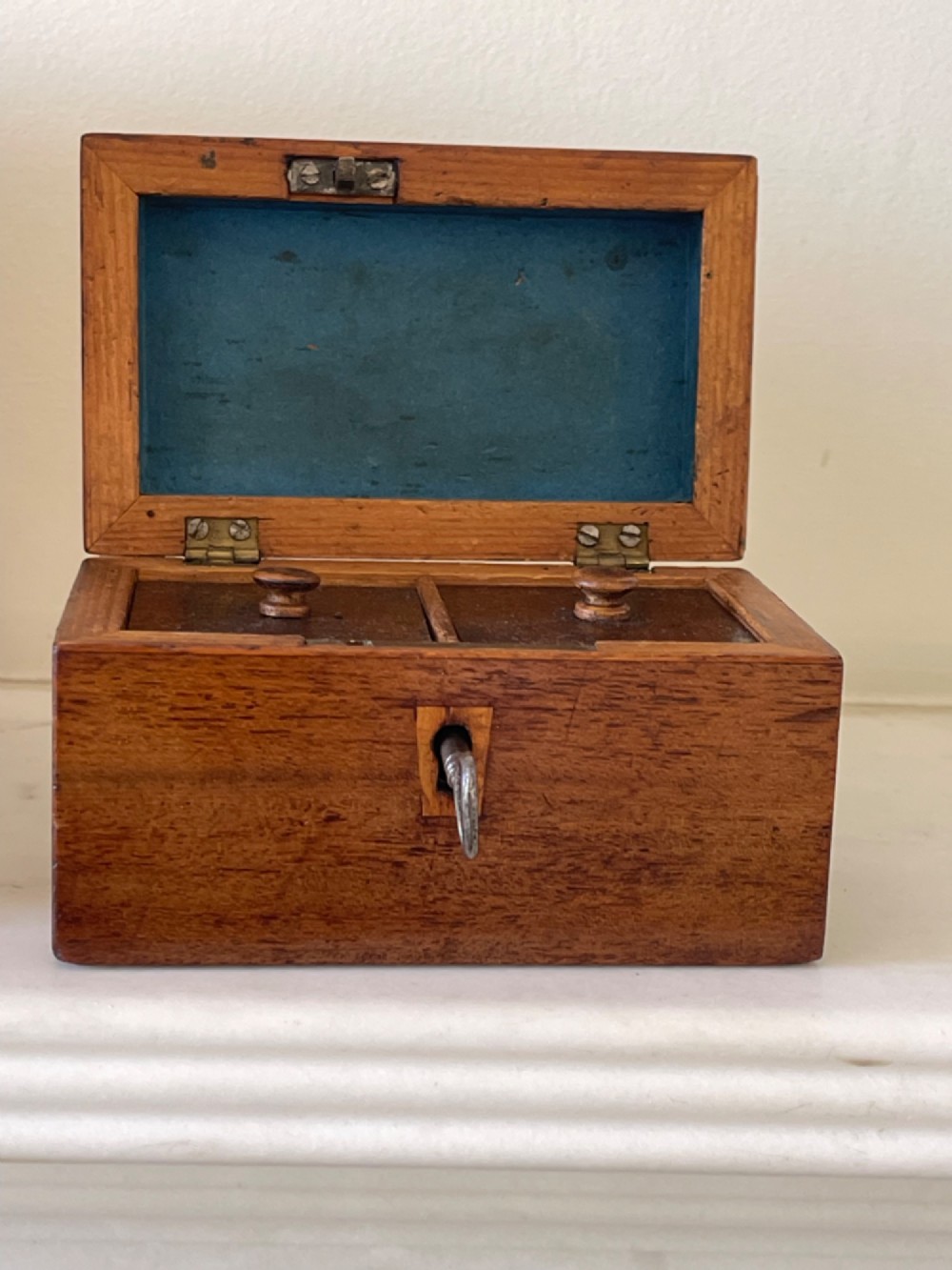 An open small wooden tea caddy featuring two internal compartments with turned knobs, a blue-lined interior lid, and a key inserted into the front lock. View V