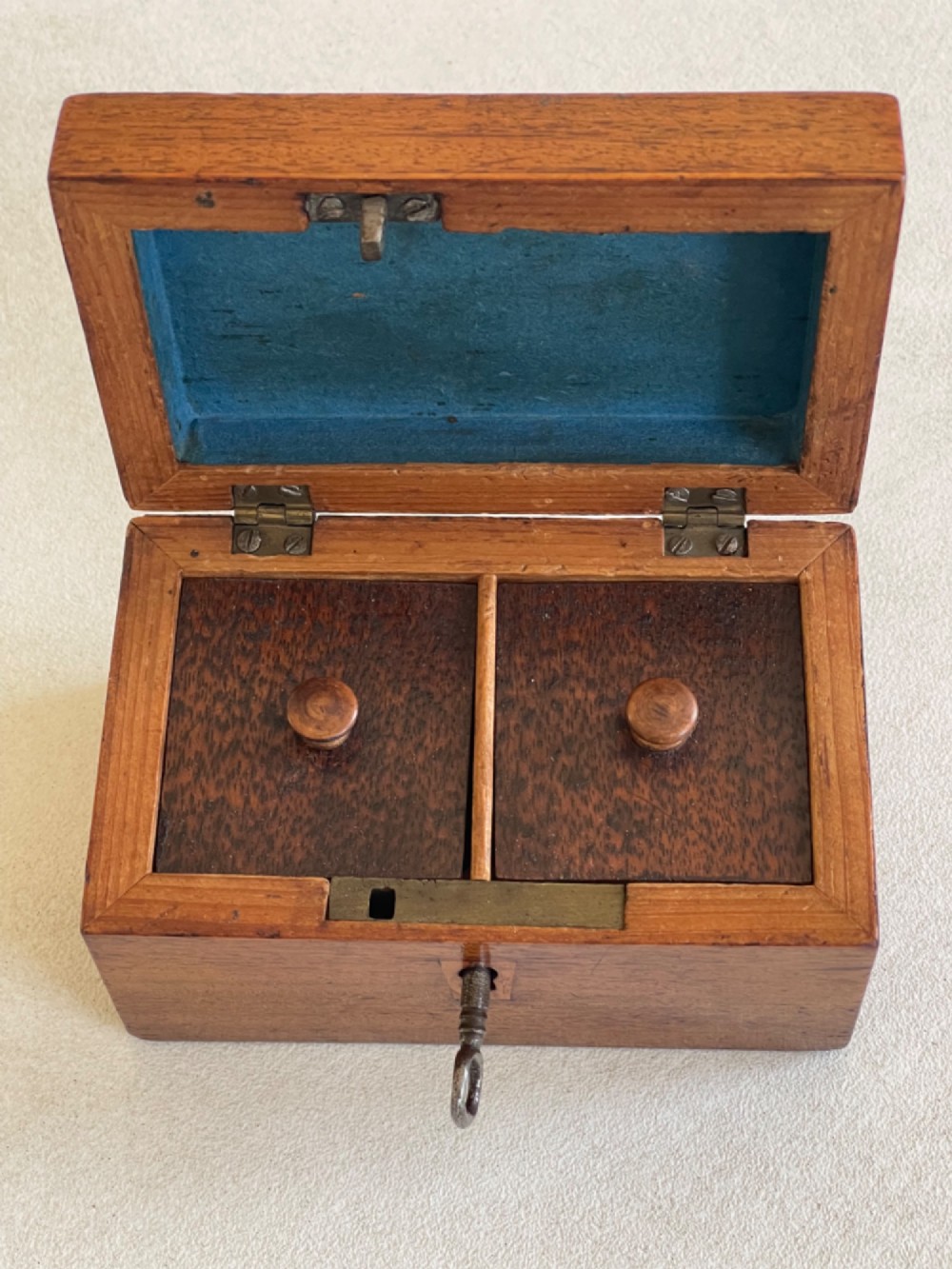 An open small wooden tea caddy featuring two internal compartments with turned knobs, a blue-lined interior lid, and a key inserted into the front lock. View VII