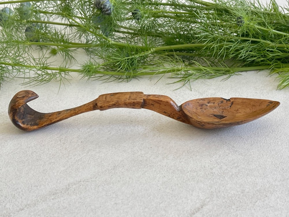A hand-carved antique wooden spoon featuring a shallow pointed bowl, a stepped shaft, and a curved terminal hook.