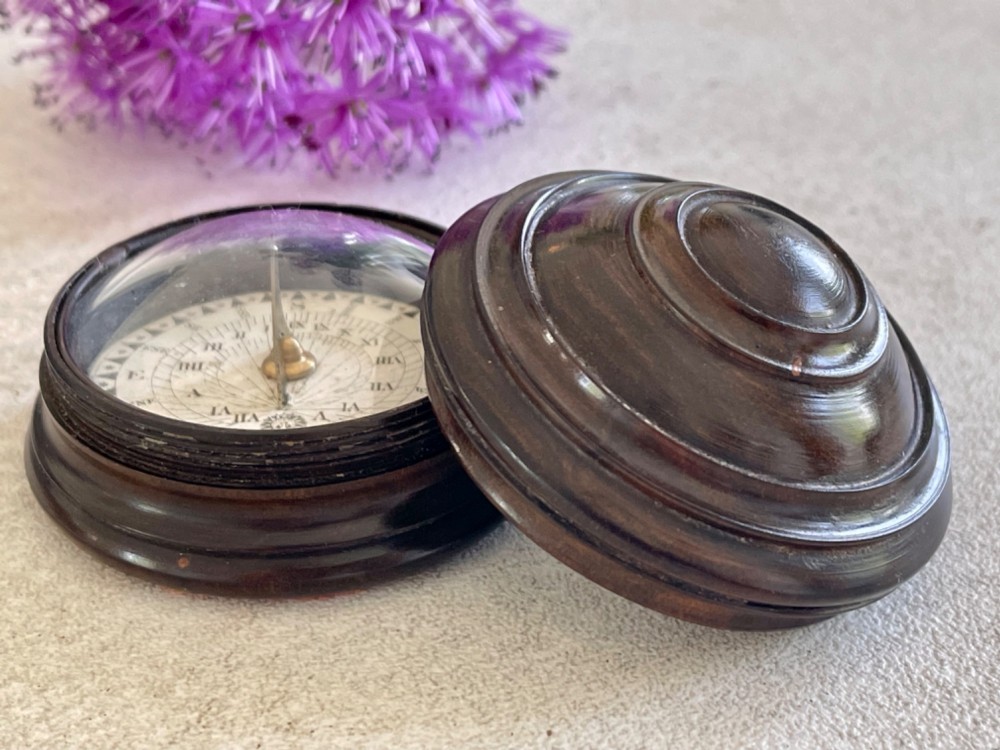 A turned dark lignum vitae wooden case opening to reveal a pocket compass and solar sundial beneath a domed glass lens.