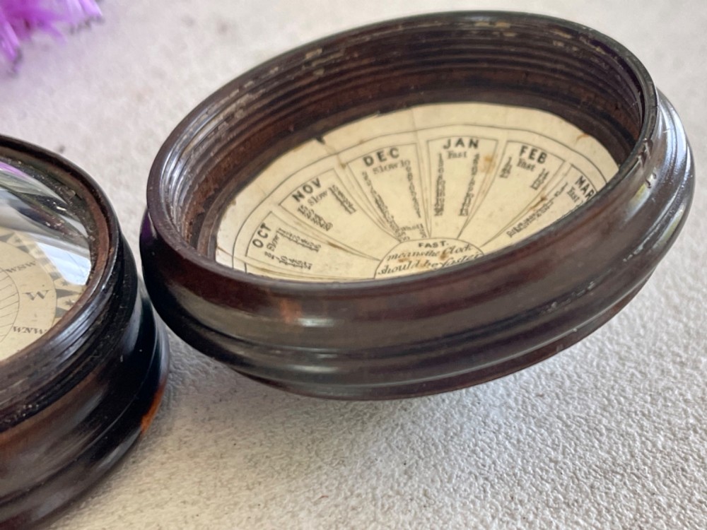 A turned dark lignum vitae wooden case opening to reveal a pocket compass and solar sundial beneath a domed glass lens. View V