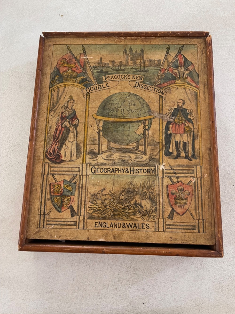 An antique wooden box featuring a printed paper label titled Peacocks New Double Dissection Geography and History England and Wales with illustrations of a globe, historical figures, and coat of arms.