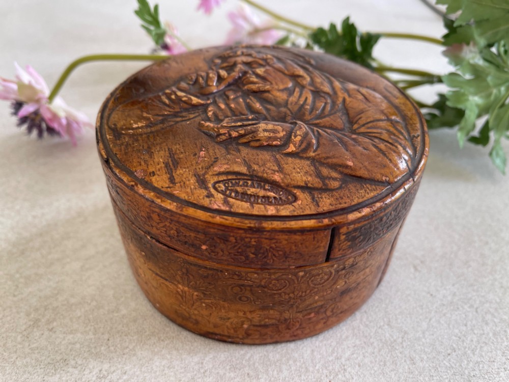 A circular brown wooden table snuff box lid decorated with a relief illustration depicting a figure taking snuff and a stamped maker mark. View II