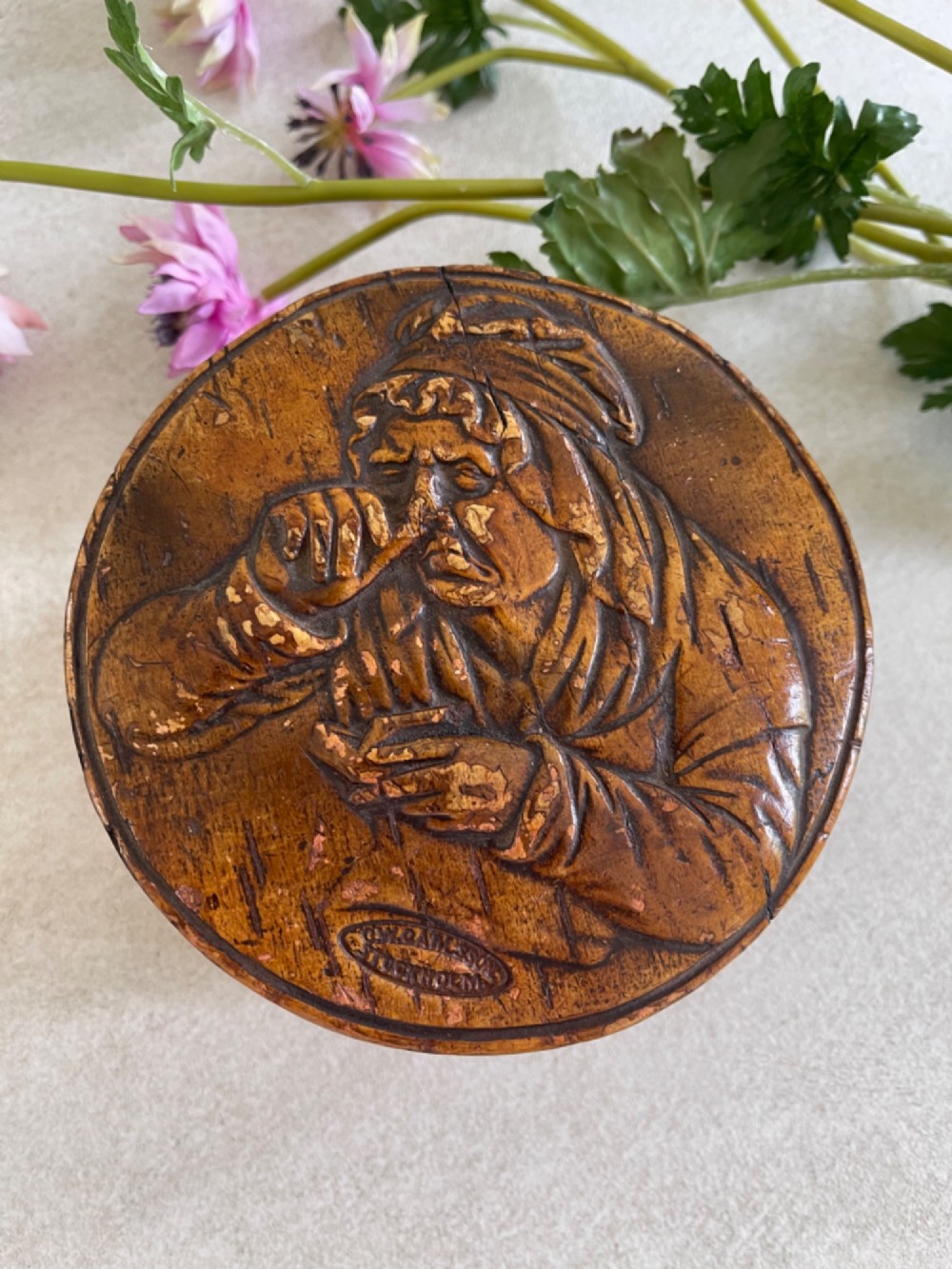 A circular brown wooden table snuff box lid decorated with a relief illustration depicting a figure taking snuff and a stamped maker mark. View III