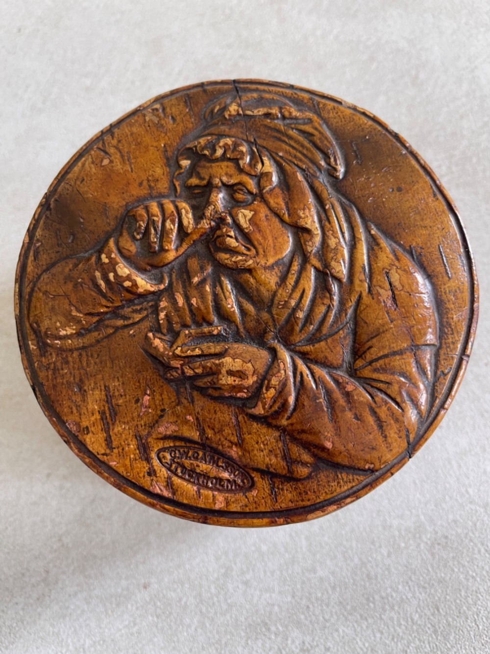A circular brown wooden table snuff box lid decorated with a relief illustration depicting a figure taking snuff and a stamped maker mark. View V