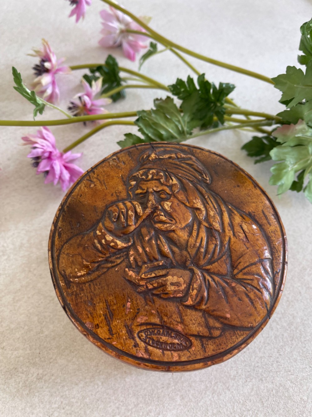 A circular brown wooden table snuff box lid decorated with a relief illustration depicting a figure taking snuff and a stamped maker mark. View VII